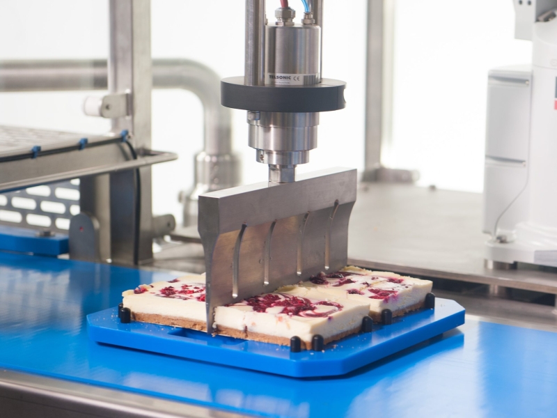 Ultrasonic Cutting Blade Cutting into Square Cheesecake on Blue Cutting board